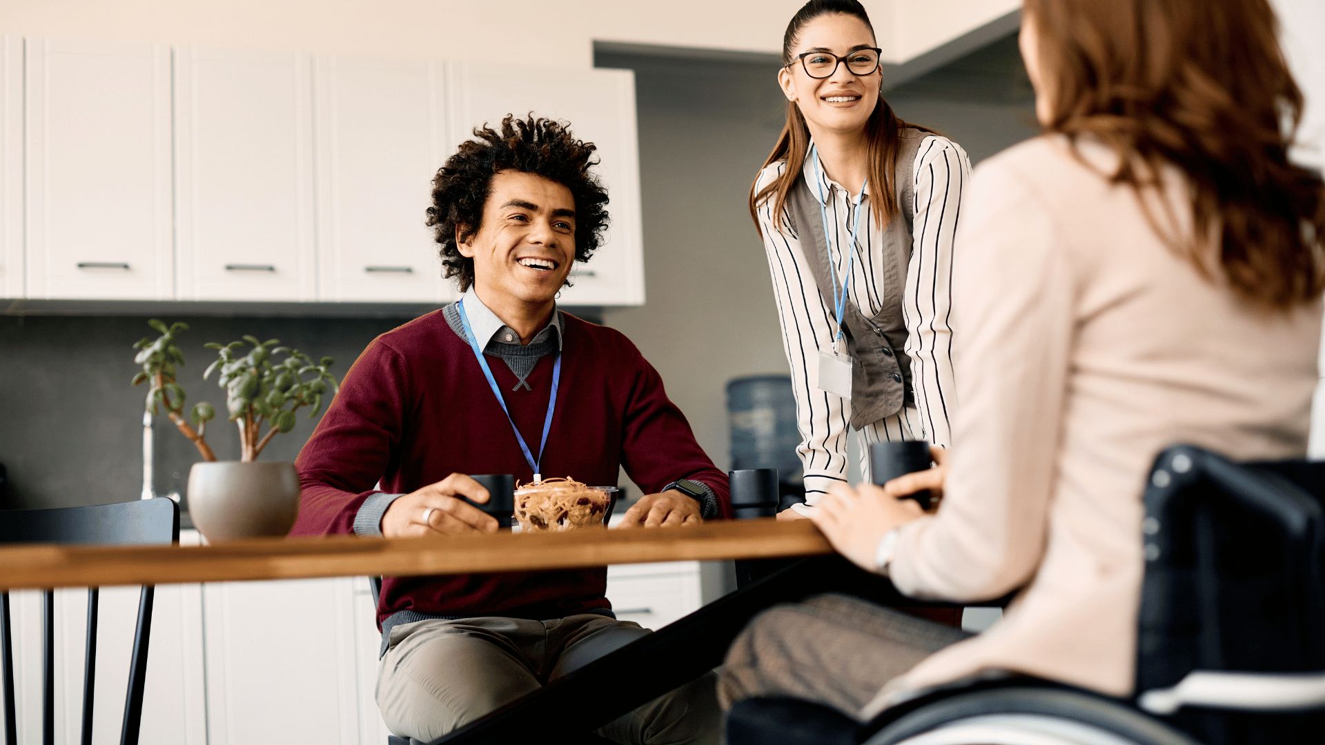 A diverse group of office workers are talking and smiling around a break room table.