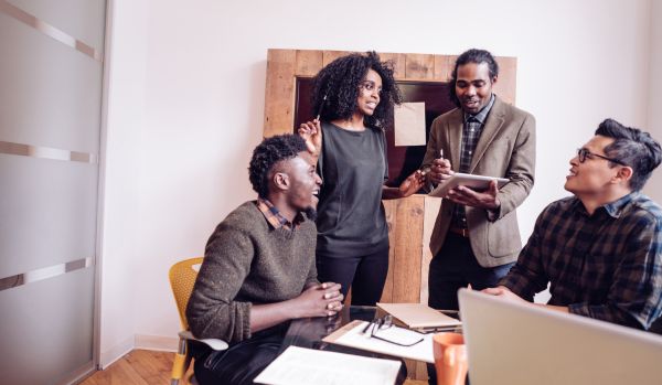 A diverse team of people are working around a desk. Some are standing, some are sitting. All are very animated and excited.