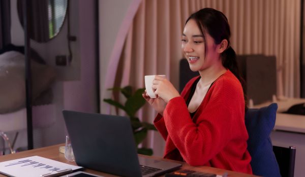 A young female-presenting remote worker is smiling at her laptop while holding a cup of coffee.