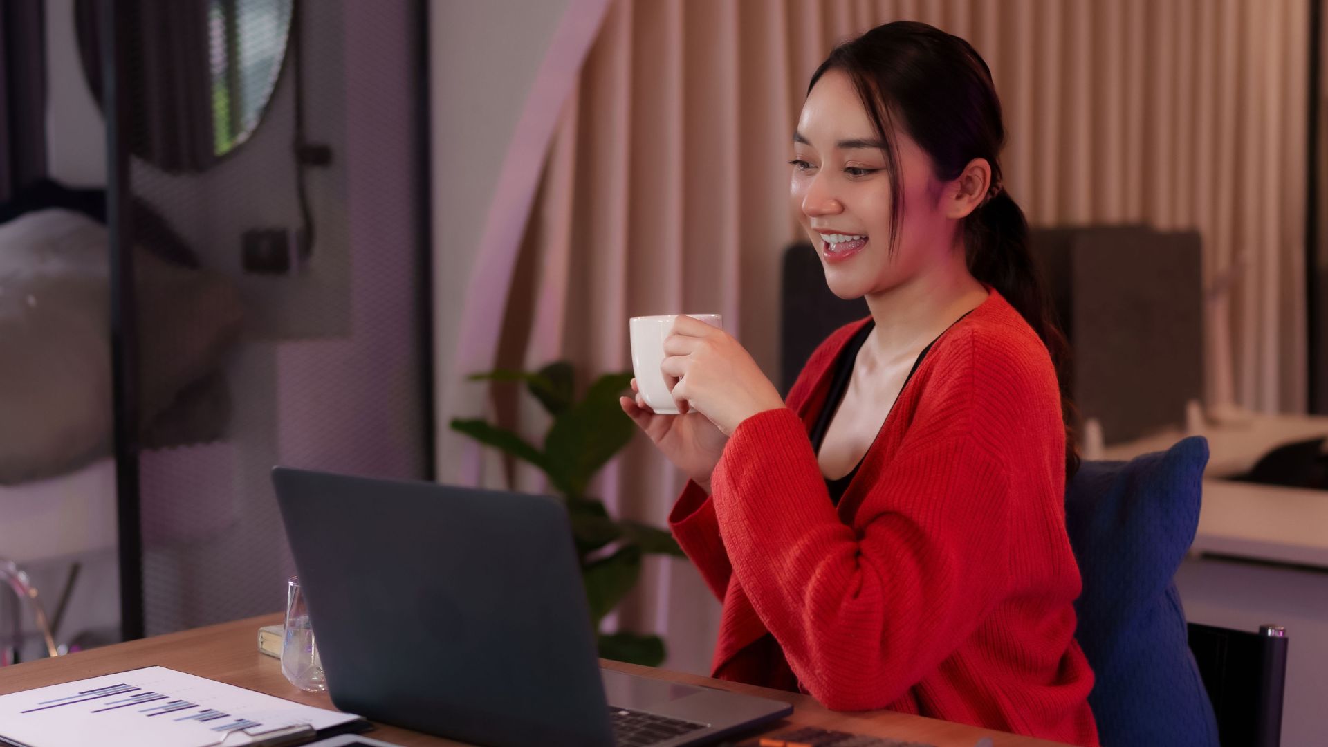 A young female-presenting remote worker is smiling at her laptop while holding a cup of coffee.