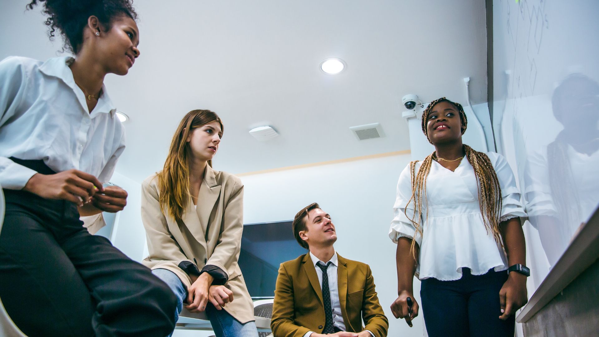 A diverse group of employees participates in a lunch & learn on effective communication. They are engaged in a discussion, with one person standing and speaking while the others listen attentively. The setting is a modern office with a whiteboard in the background.