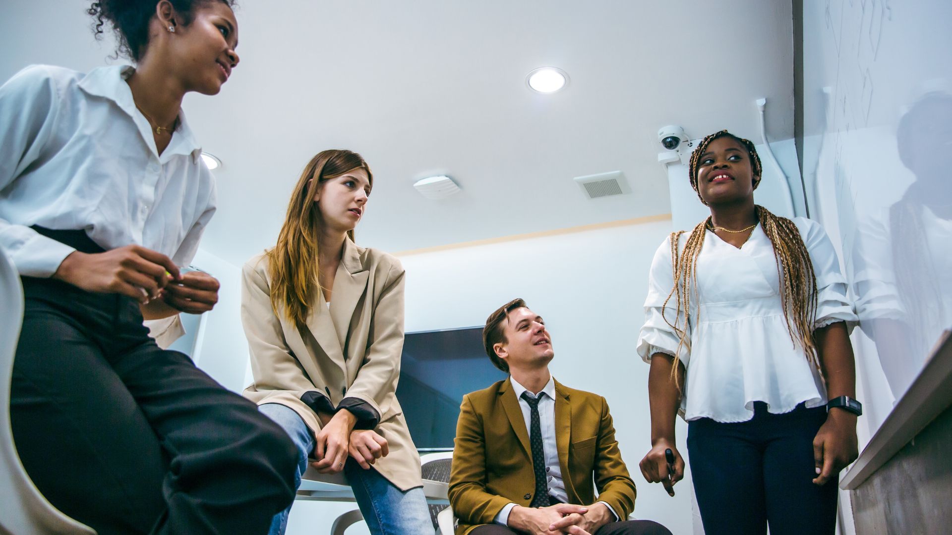 A diverse group of employees participates in a lunch & learn on effective communication. They are engaged in a discussion, with one person standing and speaking while the others listen attentively. The setting is a modern office with a whiteboard in the background.