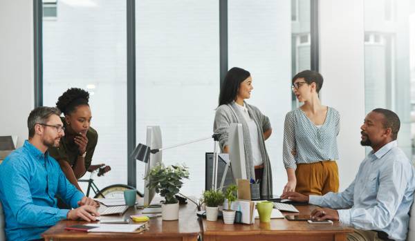 There are five employees in a conference room having a meeting at table.