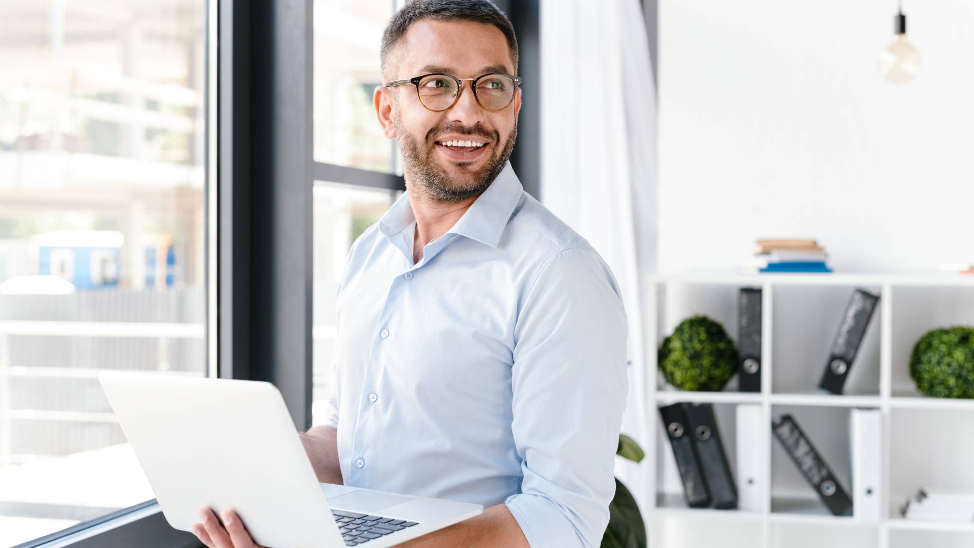 A man is standing at a window holding his laptop. He is looking over his shoulder and smiling.