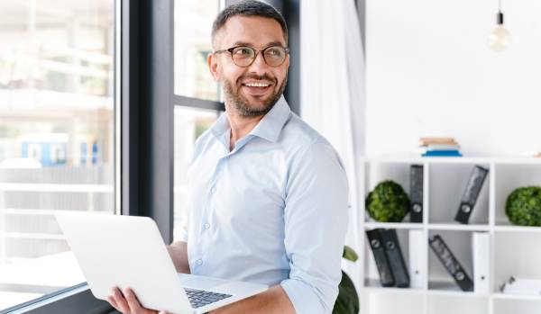 A man is standing at a window holding his laptop. He is looking over his shoulder and smiling.