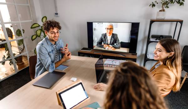 A group of employees are sitting around a conference table with another colleague on the TV via video chat.