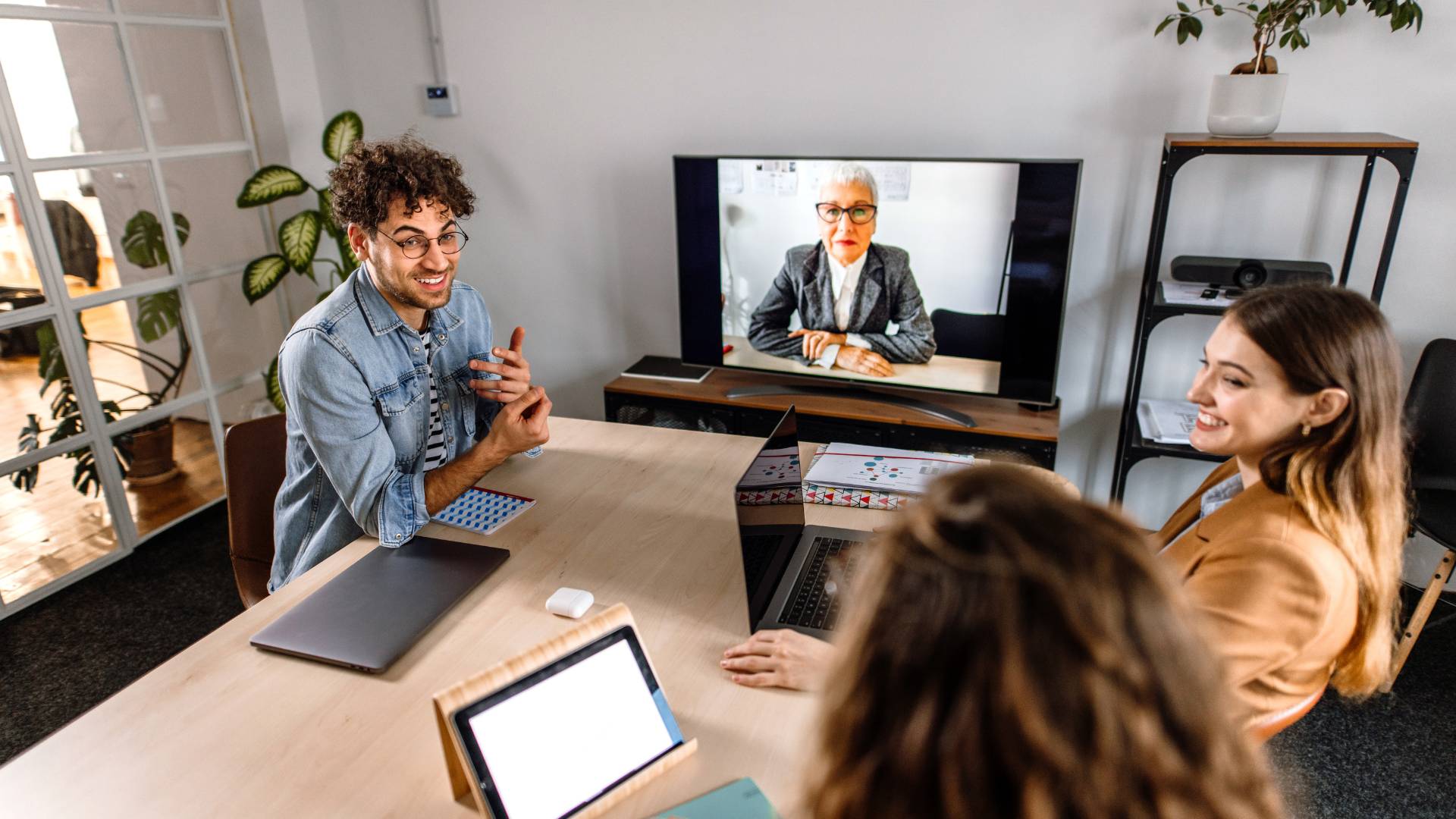 A group of employees are sitting around a conference table with another colleague on the TV via video chat.