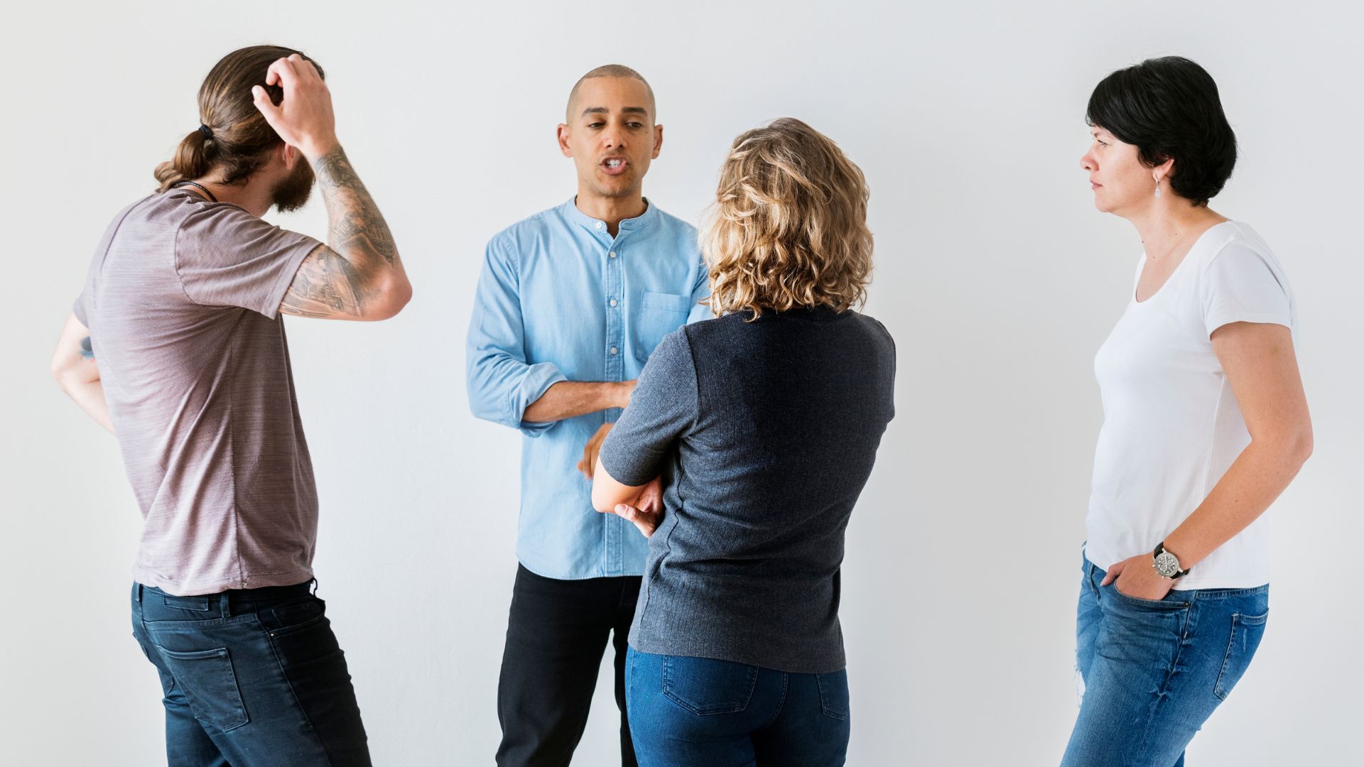A group of four adults engaged in a discussion during a training session on healthy disagreements. The individuals, two women and two men, are in a brightly lit room with a plain white background. One man gestures while speaking to the group, one woman listens intently, another man appears stressed and is running his hand through his hair, and the other woman observes the interaction.