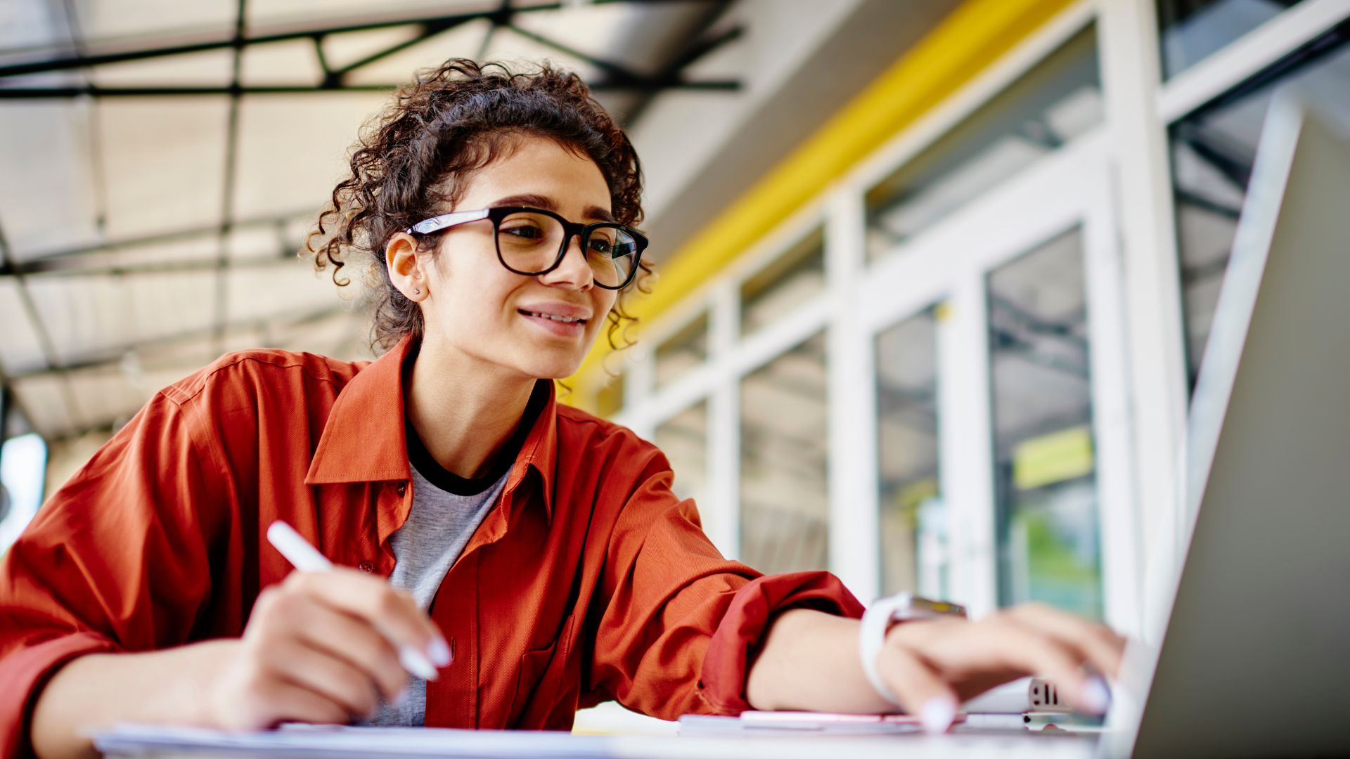 A young woman wearing glasses is working outside a cafe. She has one hand on her laptop and another writing in a notebook as she prioritizes her work
