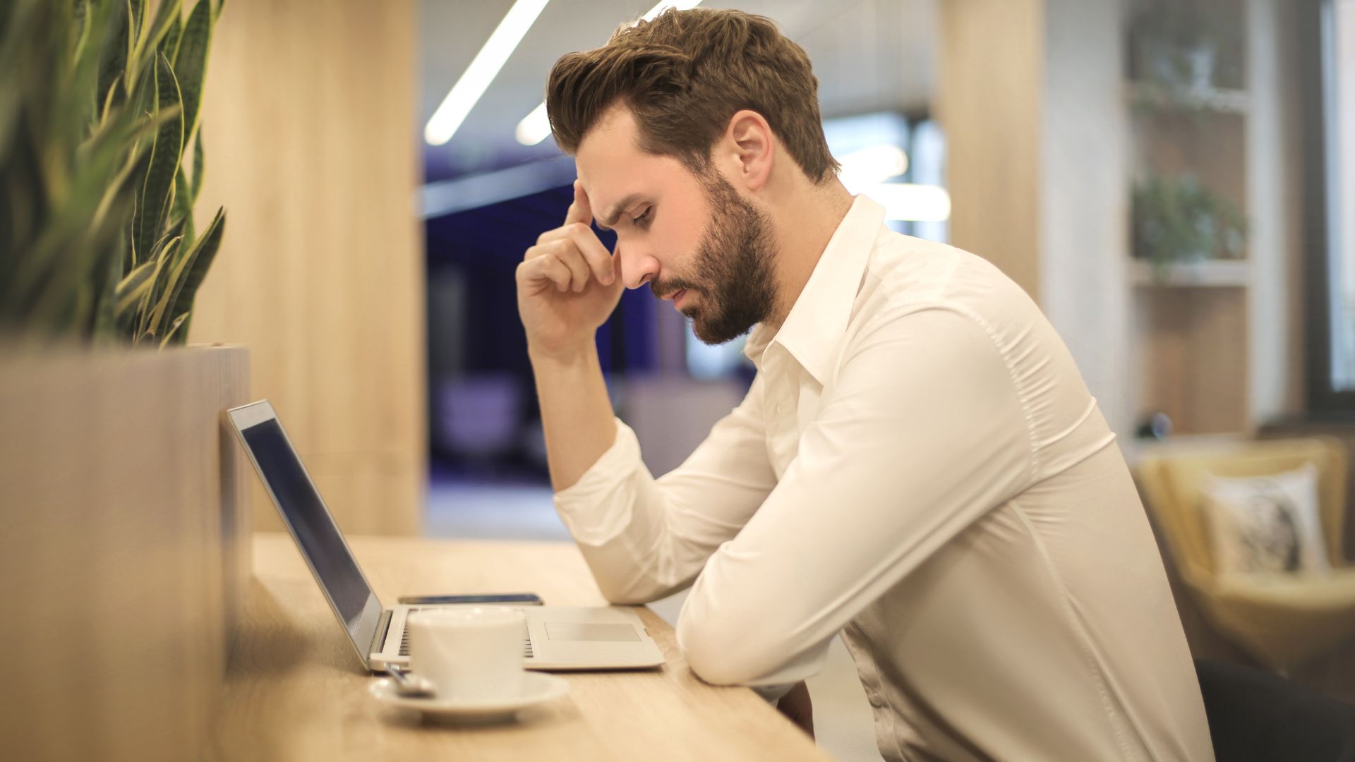 A young bearded man is thinking and looking at something on his laptop. We're looking at him from the side.