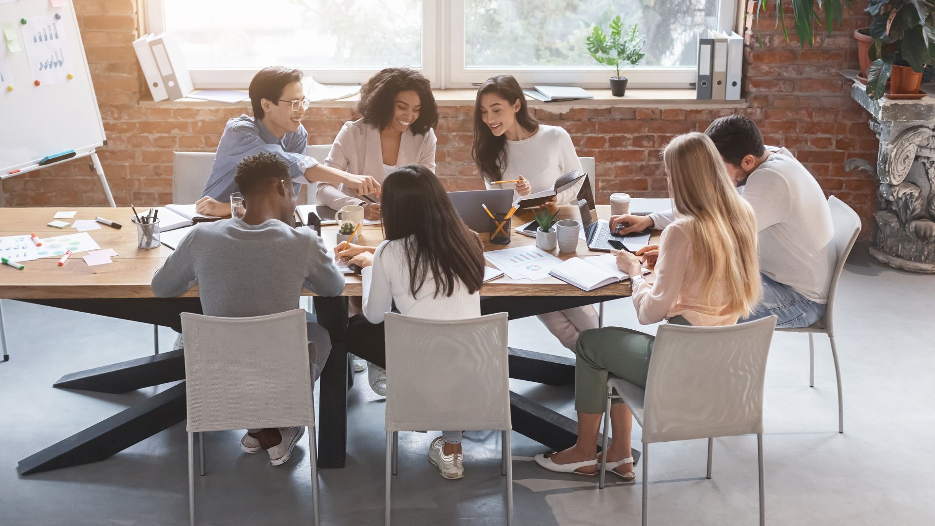 A diverse group of employees are working together on a project around a conference table with a window in the background.