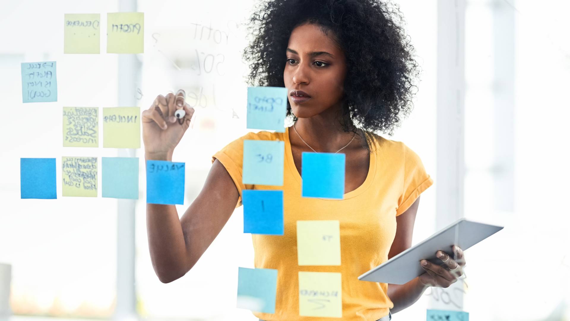 A woman is standing in front of a transparent board that has blue and yellow sticky notes on it. She is holding a tablet in one hand and she is writing on the glass board with the other.