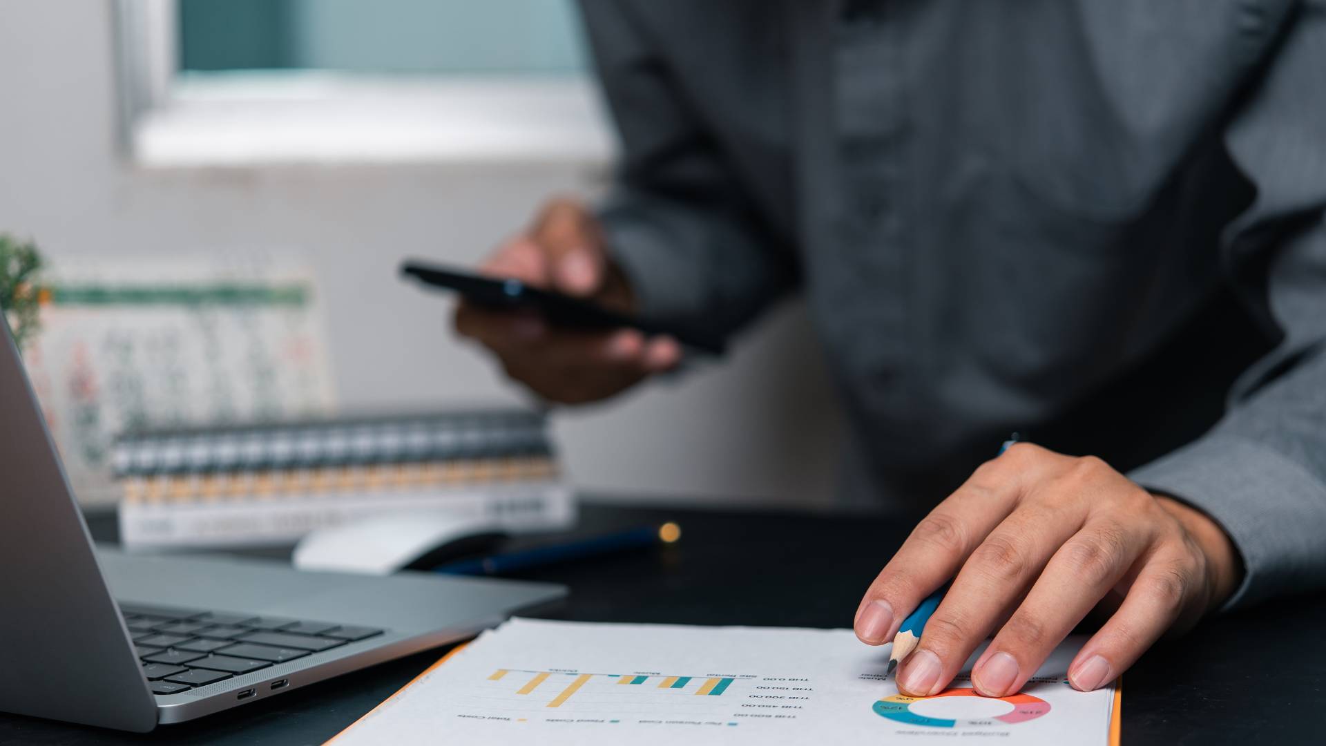 A man is reviewing some financial reports at his laptop with his phone in his hand. We can only see the man from the shoulders down.