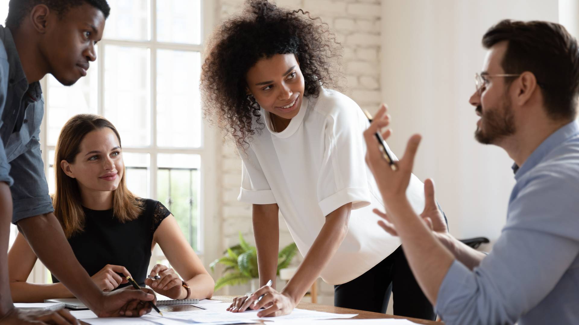 There are four people who are in a meeting. One woman is sitting down at the table along with a man who is also sitting. There is a woman and another man who are standing and engaging with the other two people.