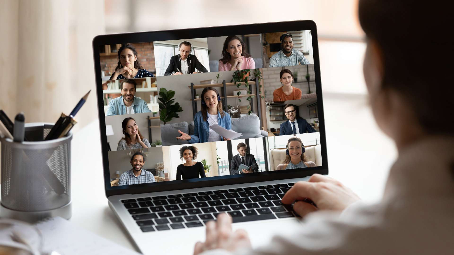 We are looking over a man's shoulder at his laptop on his desk. He is on a virtual training with a diverse group of people.