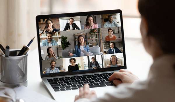 We are looking over a man's shoulder at his laptop on his desk. He is on a virtual training with a diverse group of people.