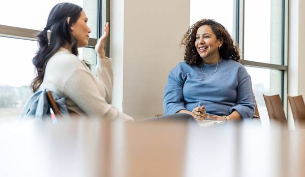 Two women sitting in an office having an engaing conversation. One woman is wearing a white shirt and the other is wearing a blue shirt.