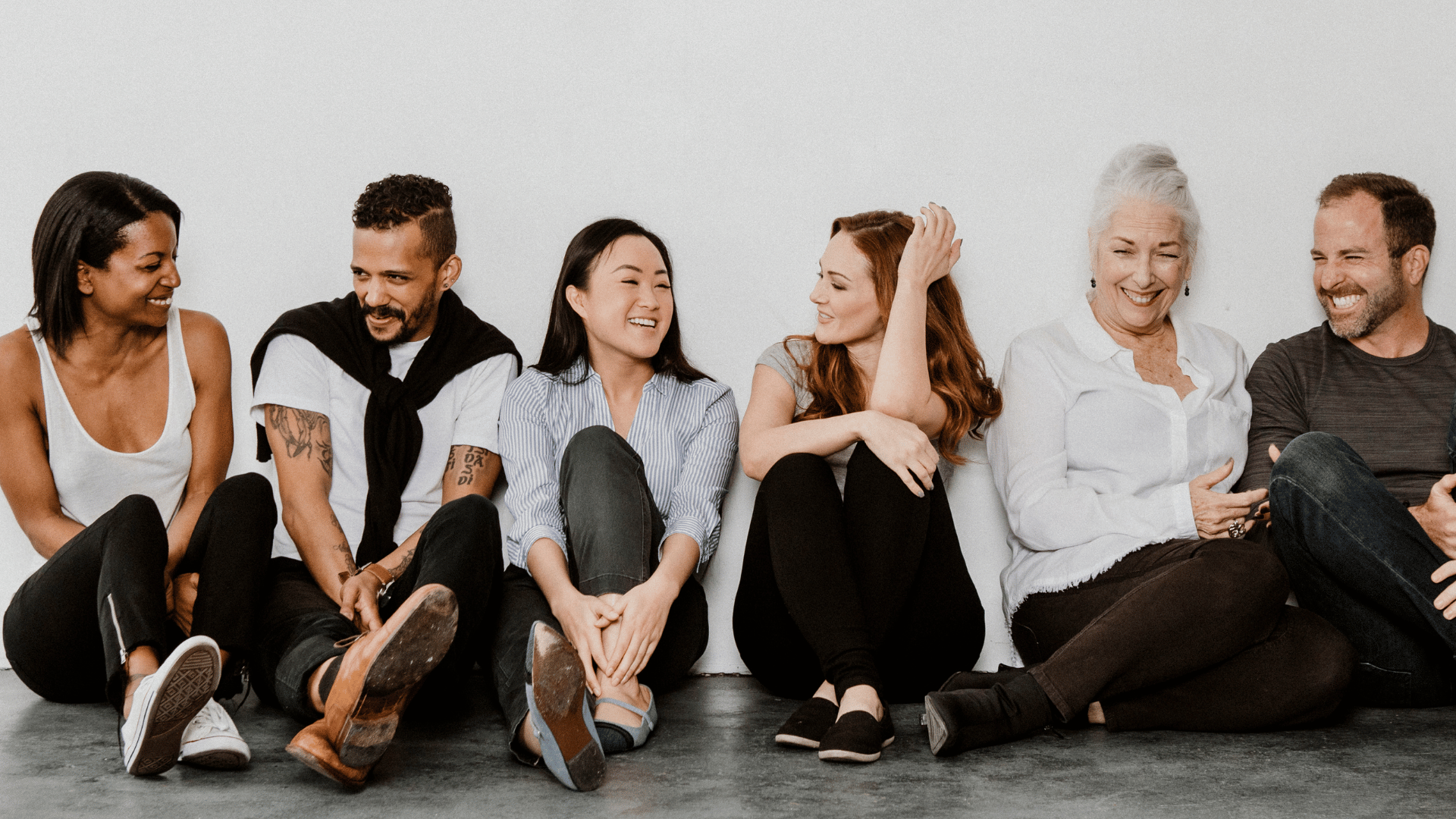 Group of people sitting and chatting while leaning against a white wall.