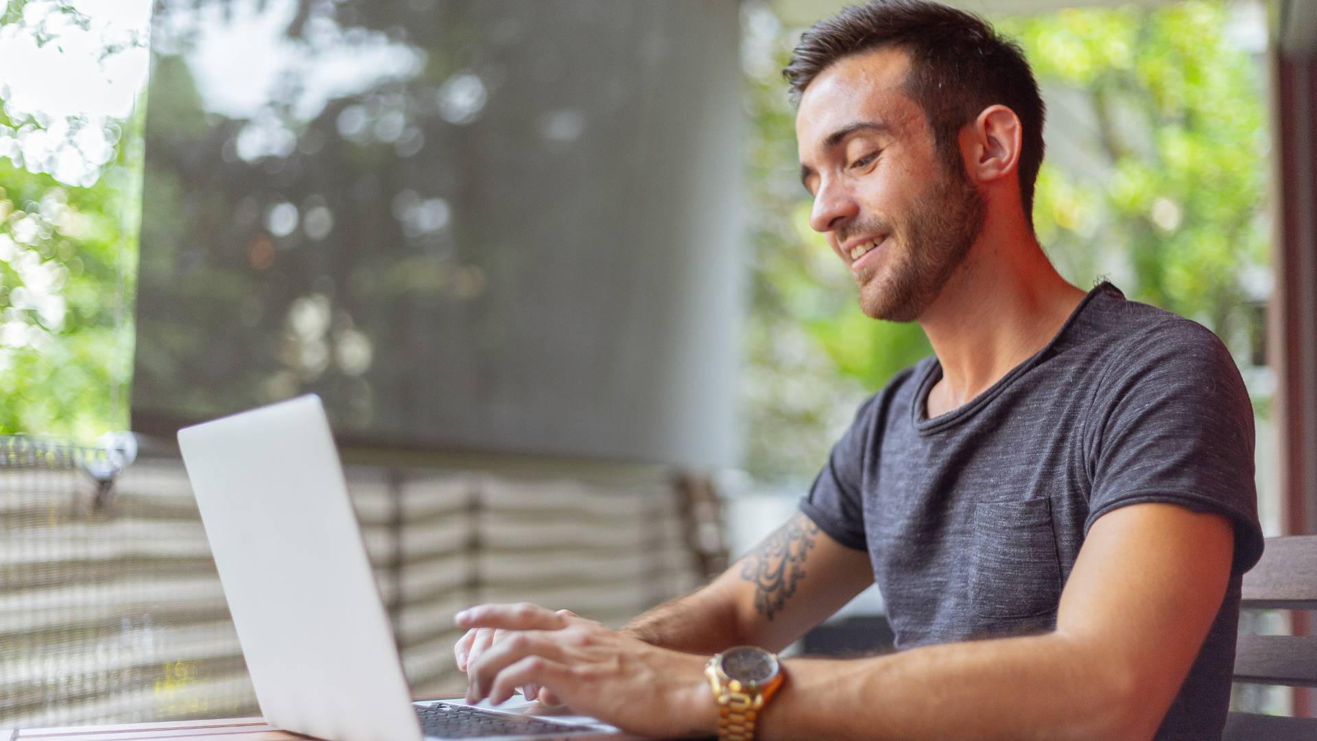 A young man in a short-sleeve t-shirt is working on his laptop outside on a patio.