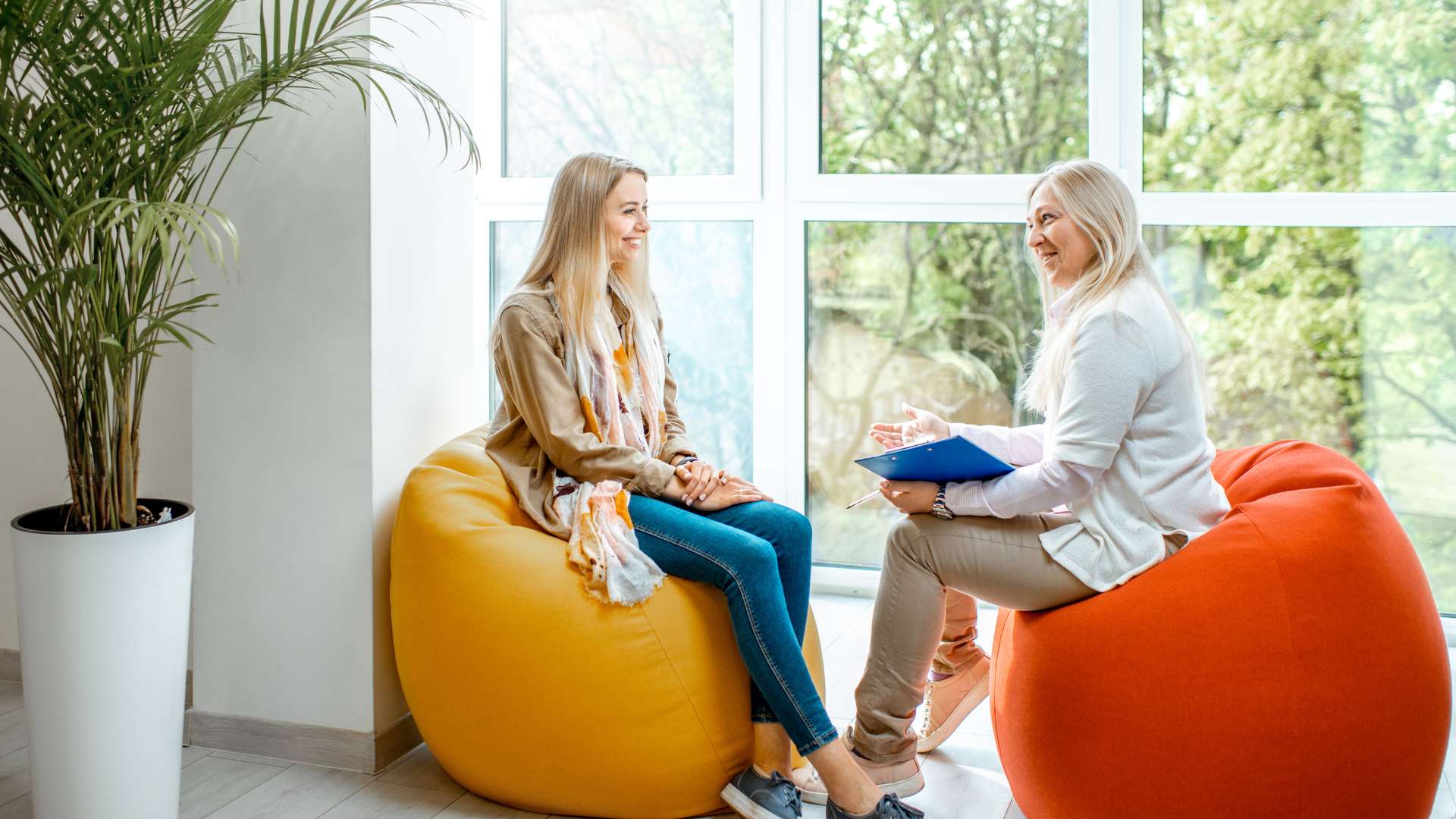 Two women have a conversation in an office next to a window, sitting on bean bag chairs.