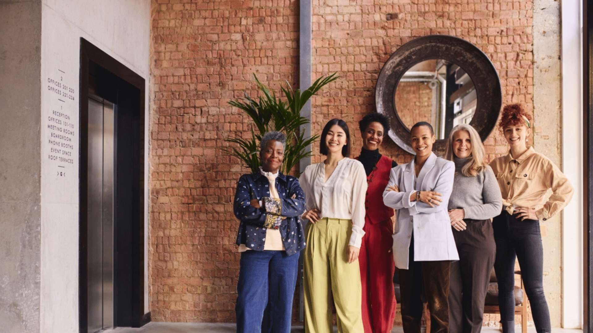 A group of diverse women are standing together in front of a brick wall in an office.