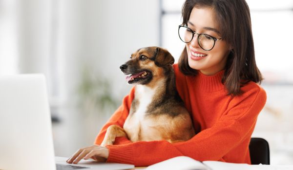 A young woman wearing glasses and an orange sweater is working on her laptop with her small dog sitting in her lap. Both are smiling.