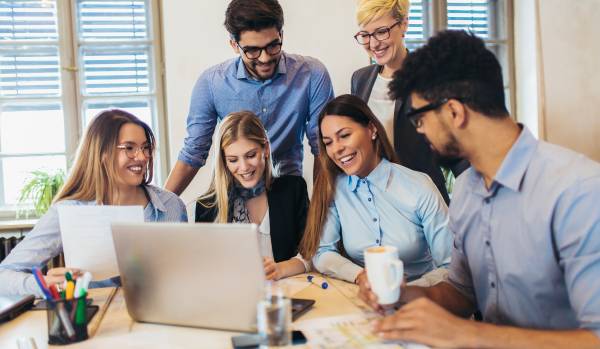 There are six people gathered around a table in an office laughing, smiling, and talking. Four of them are sitting at the table and two are behind them standing. There's a laptop on the table.
