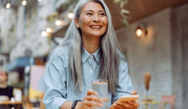 An Asian woman with long gray hair is smiling and holding a glass of water while sitting at an outdoor table.