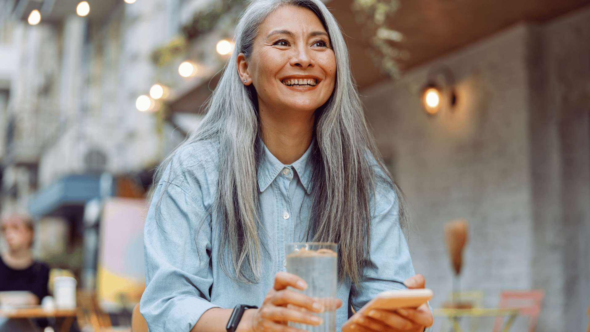 An Asian woman with long gray hair is smiling and holding a glass of water while sitting at an outdoor table.