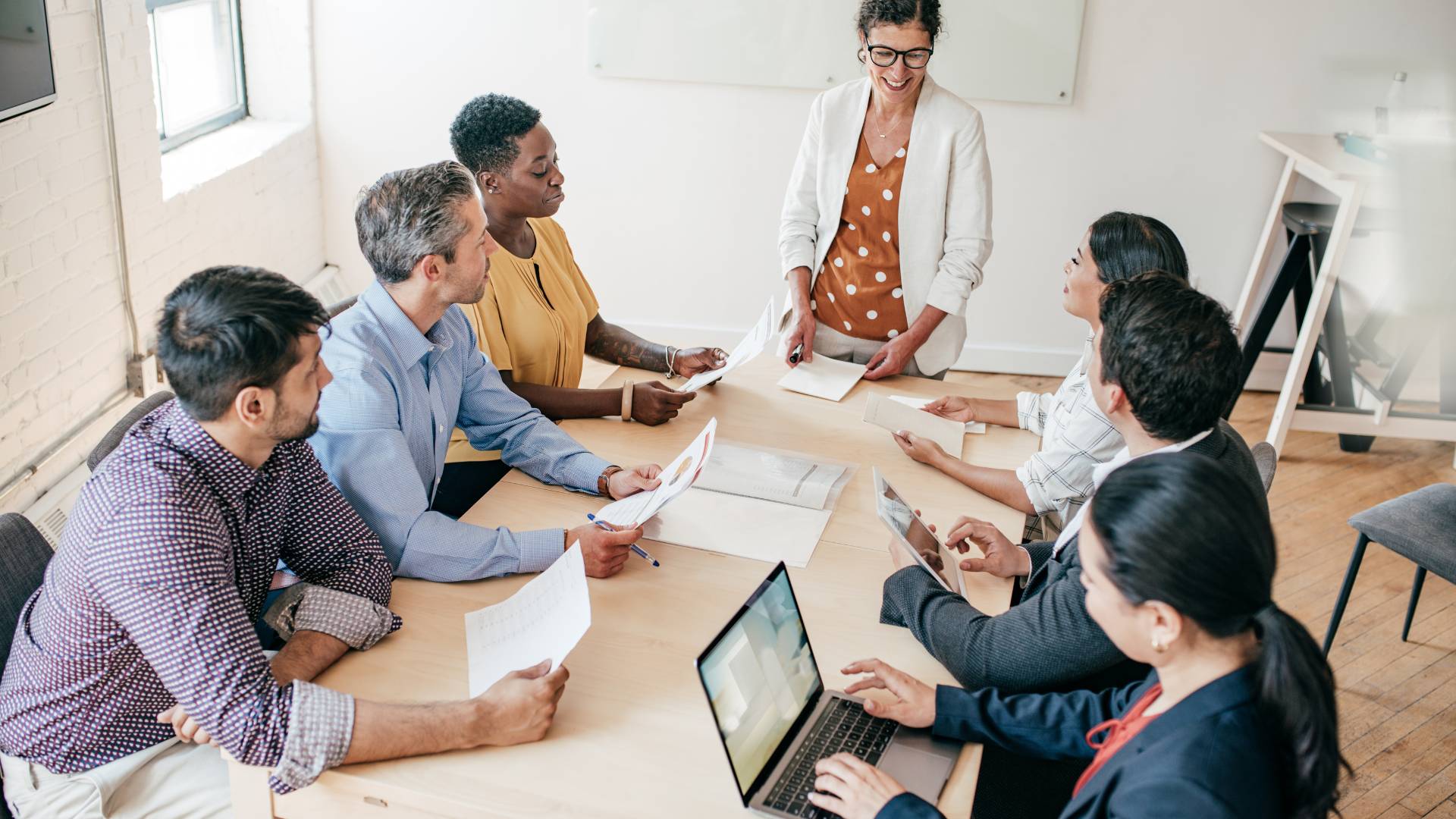 We are looking down at a team sitting at a conference table. A woman is standing st the far end leading a discussion.