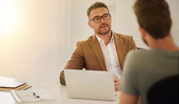 A man is having a feedback conversation with a member of his team. We are looking over the team member's shoulder at the man's face.