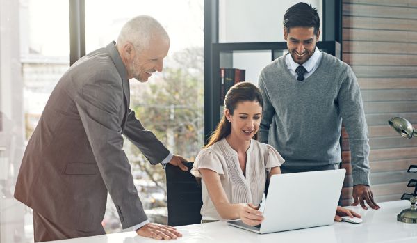 A woman is sitting at a desk in front of a computer. Behind her are two men looking over her shoulder at her computer giving her feedback. They are all happy.