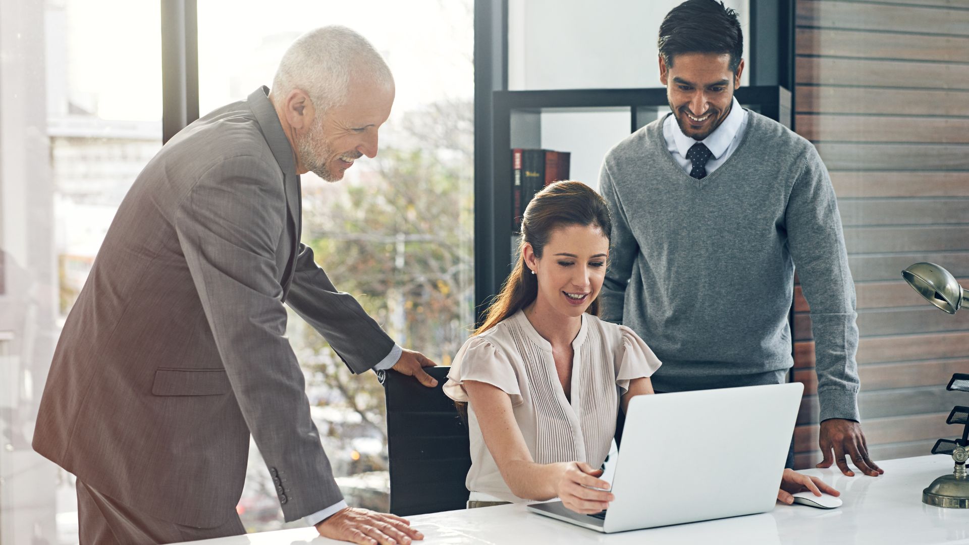 A woman is sitting at a desk in front of a computer. Behind her are two men looking over her shoulder at her computer giving her feedback. They are all happy.