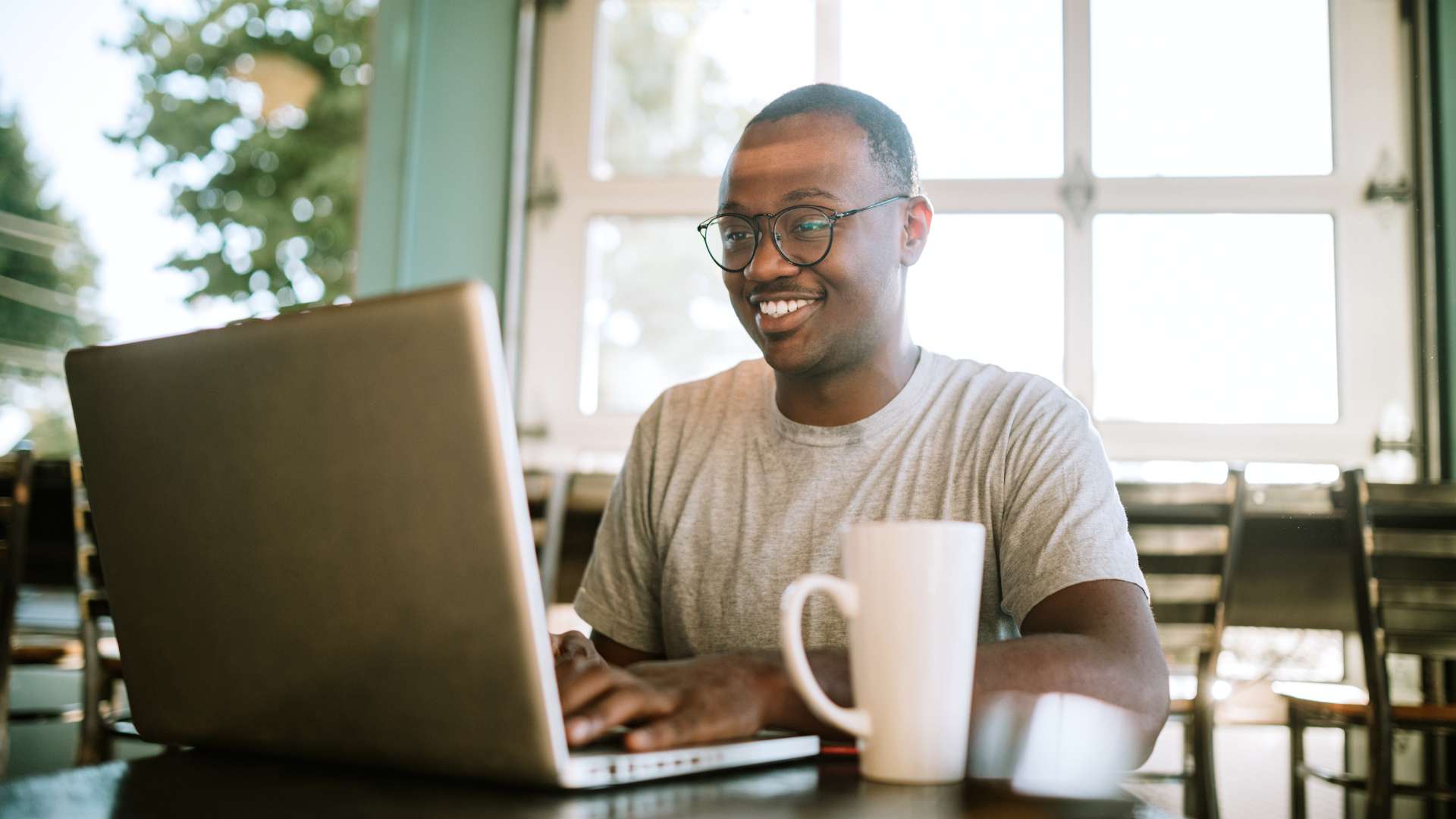 A young man with glasses works on his laptop from a coffee shop, smiling.