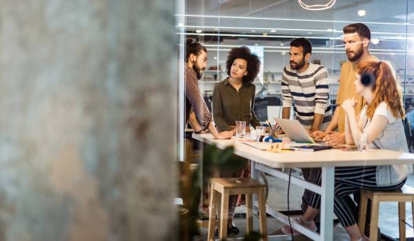 There are five team members standing around a table in an office having a meeting. One is a woman and she is sitting down and wearing a pair of black headphones. The office has an industrial feel to it.