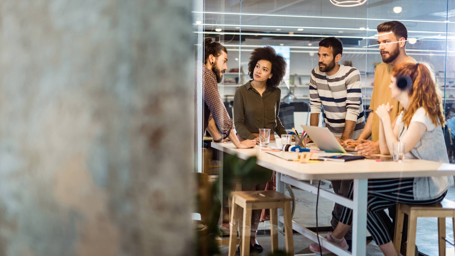 There are five team members standing around a table in an office having a meeting. One is a woman and she is sitting down and wearing a pair of black headphones. The office has an industrial feel to it.