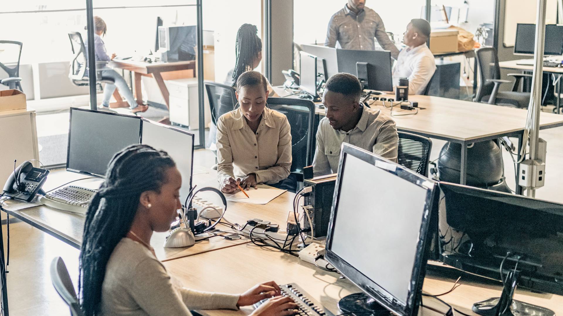 There are seven people in a large office. Three are working at a communal table which have computers on them. Another three are working at a different table and conversing. One woman is sitting by herself in another room at a desk working.