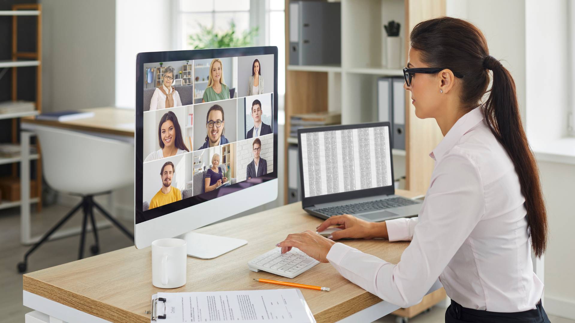 Woman sitting at desk in home office in a virtual meeting on her computer. There are nine people on the screen in the virtual meeting.