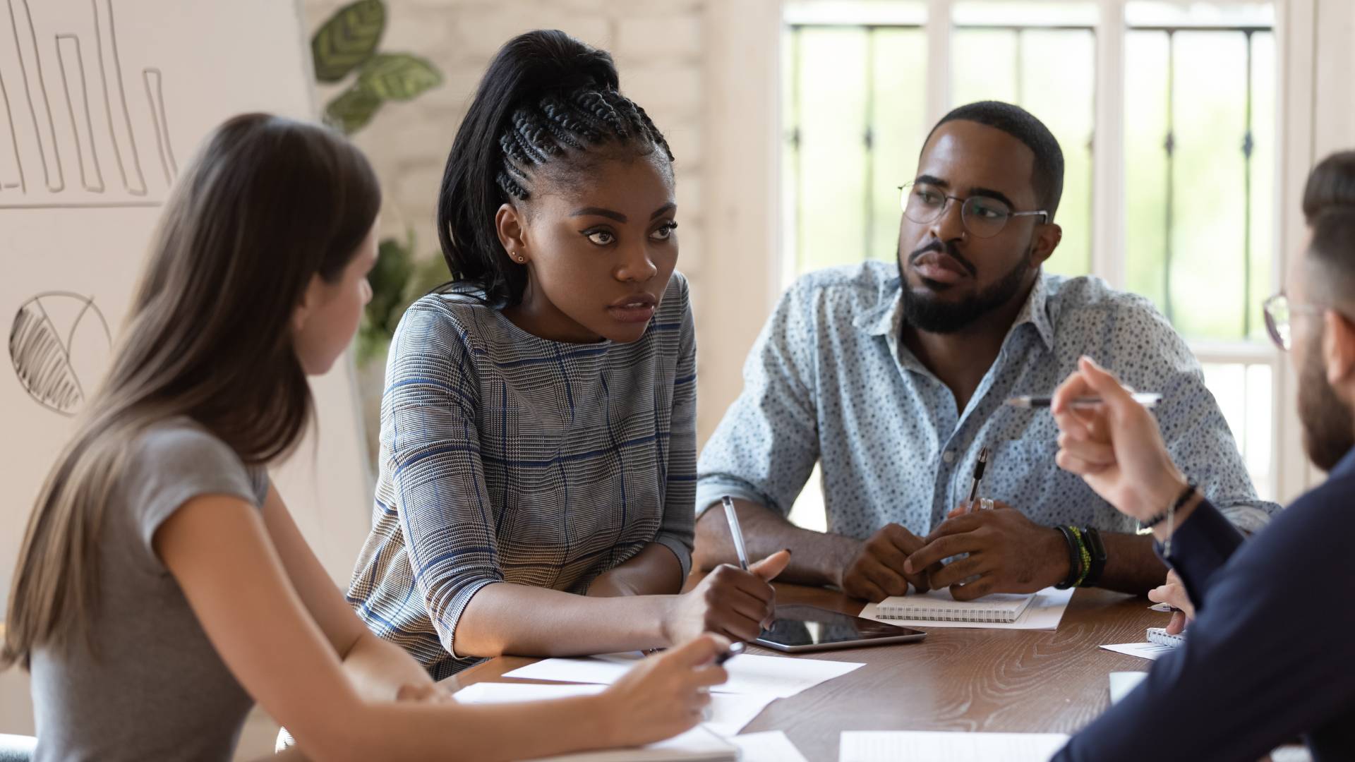 There are three employees gathered around a table in a meeting. Two are women and one is a man. There's a graph on an easel behind them.