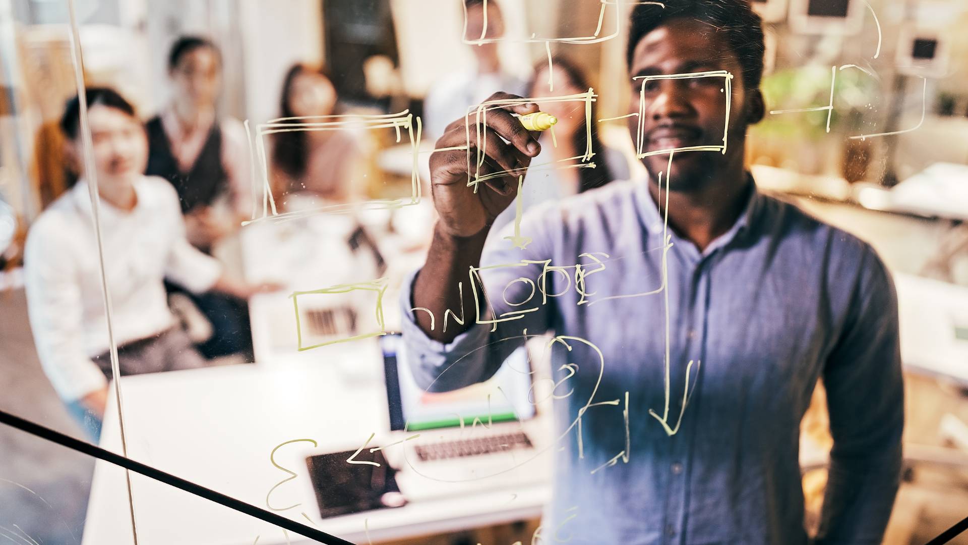 A young manager is writing on a transparent whiteboard as he teaches his team, who is sitting at a conference table behind him. We are looking through the transparent whiteboard at the man and his team.