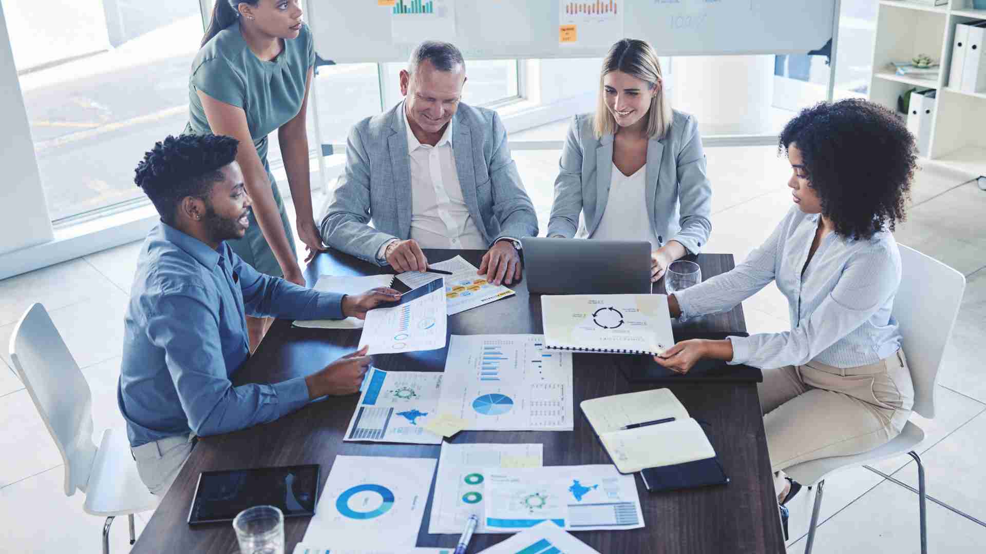 There are five managers having a meeting in a conference room. Four of them are sitting at a table while one is standing. There are documents featuring charts and graphs on the table.