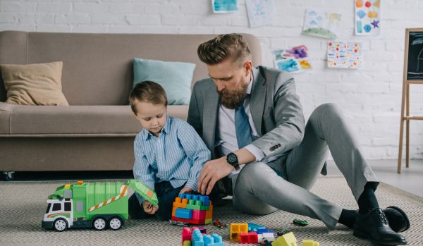 A man in a suit is sitting on the floor of his living room playing with his child.