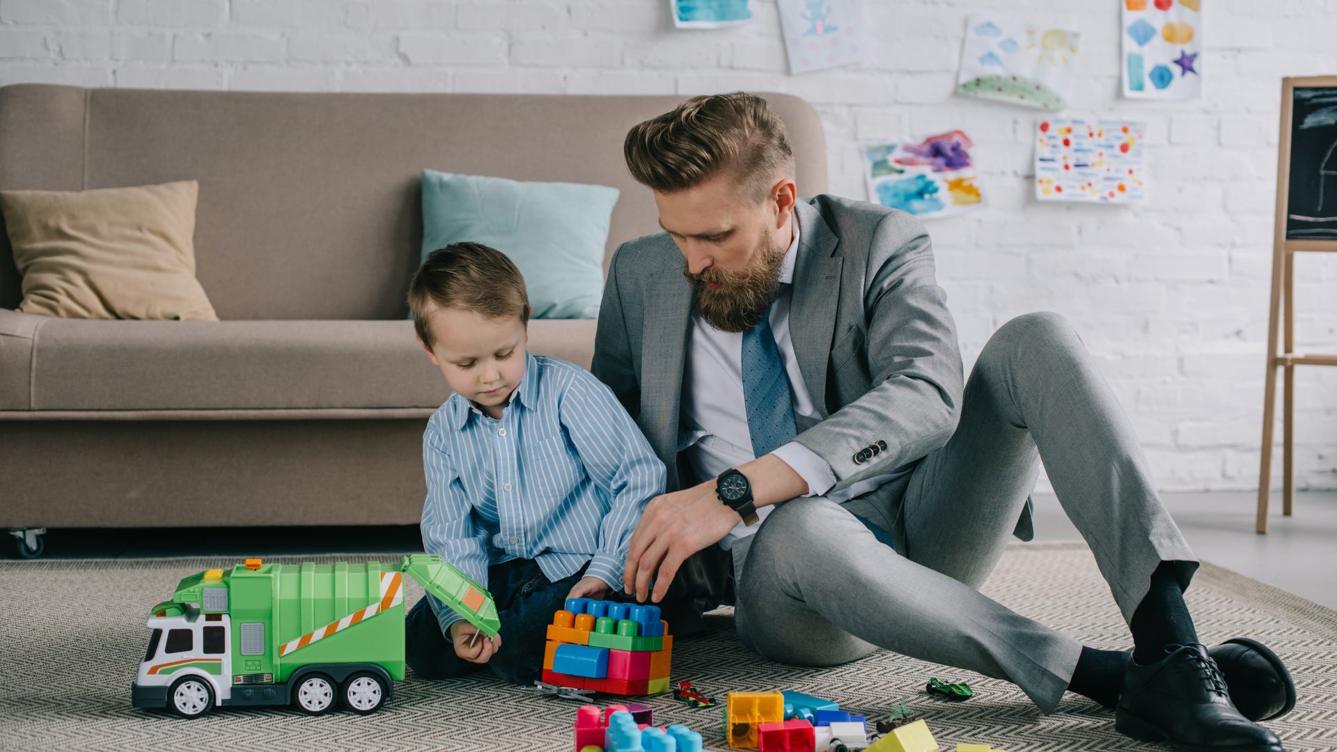 A man in a suit is sitting on the floor of his living room playing with his child.