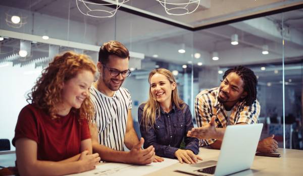 There are four people standing around a desk. They are engaged in a happy conversation and there is also a laptop on the table.