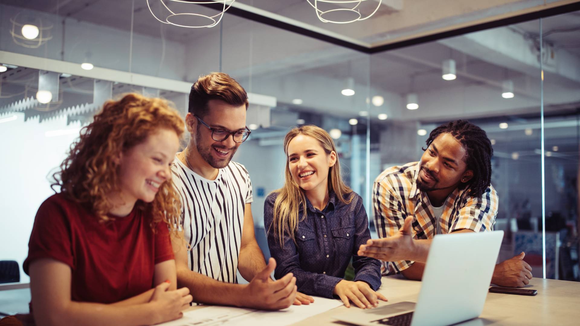 There are four people standing around a desk. They are engaged in a happy conversation and there is also a laptop on the table.
