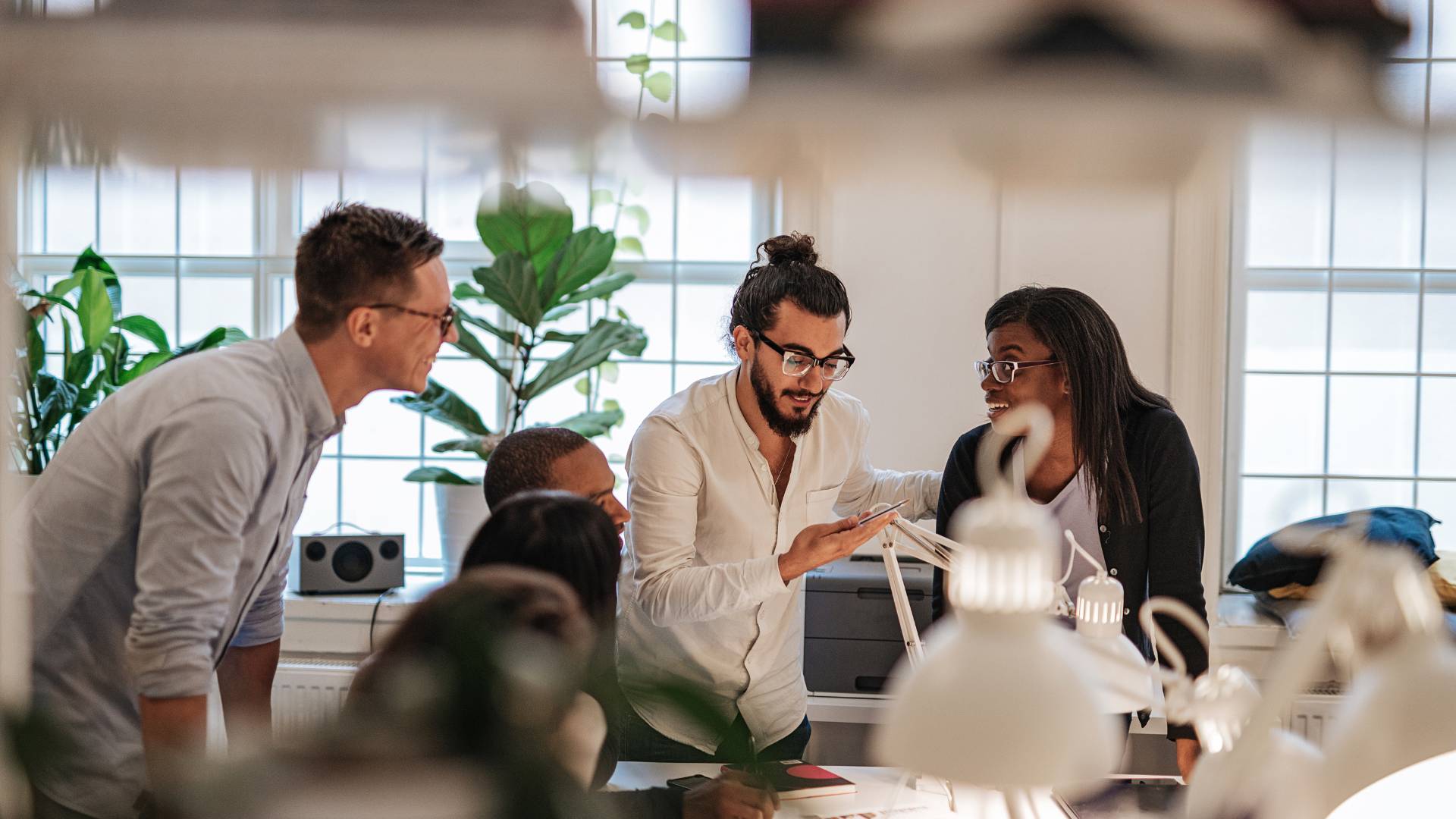 There are a group of people conversing in an office. Three are standing and the other three are sitting at a table.