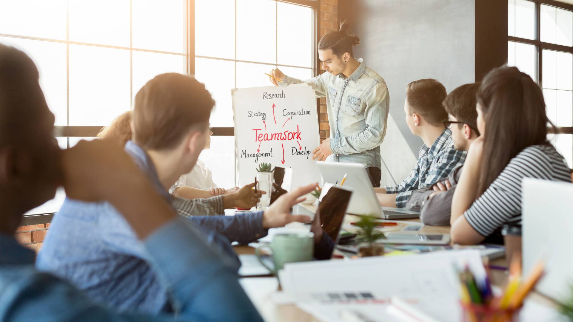 A young adult man is standing next to an easel conducting a training on teamwork. There are seven people sitting around a table watching the presentation.