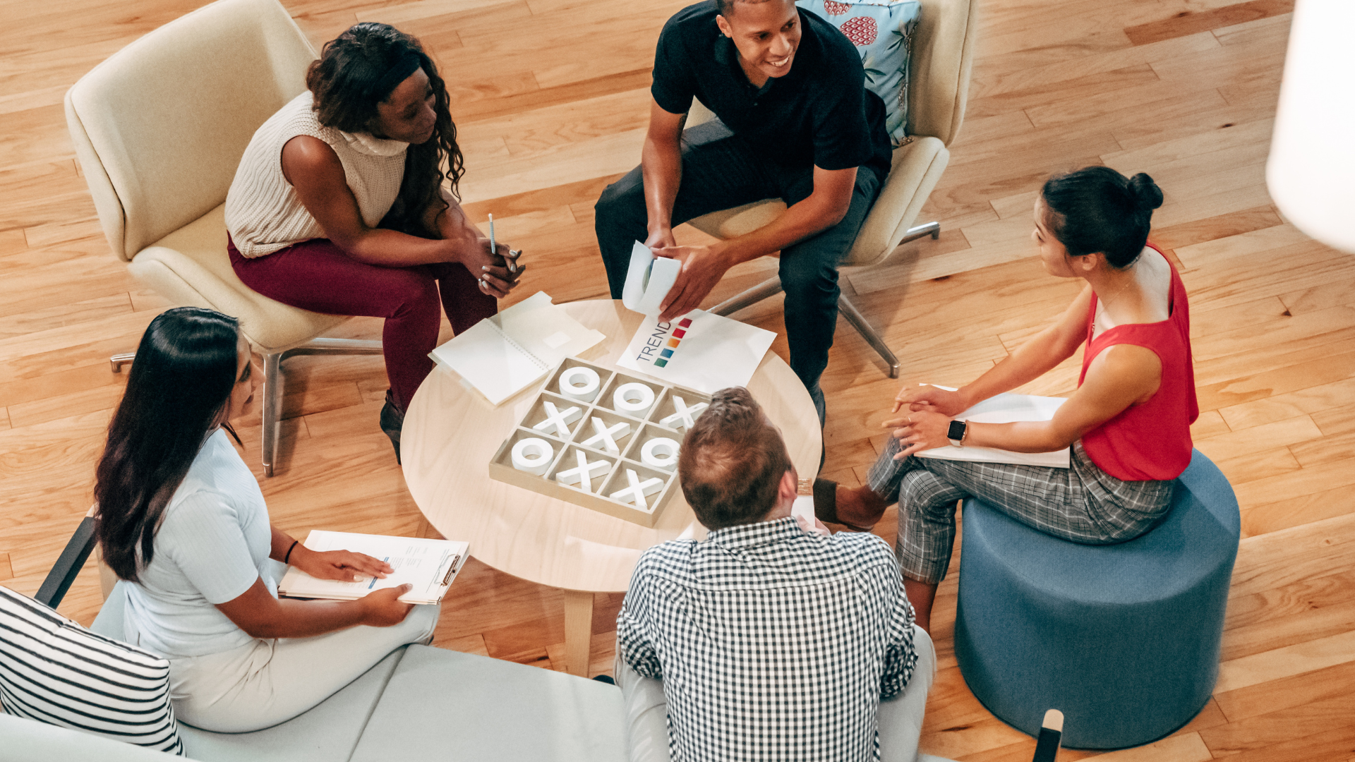 A diverse team of 5 people are sitting in a circle in a common area around a coffee table.