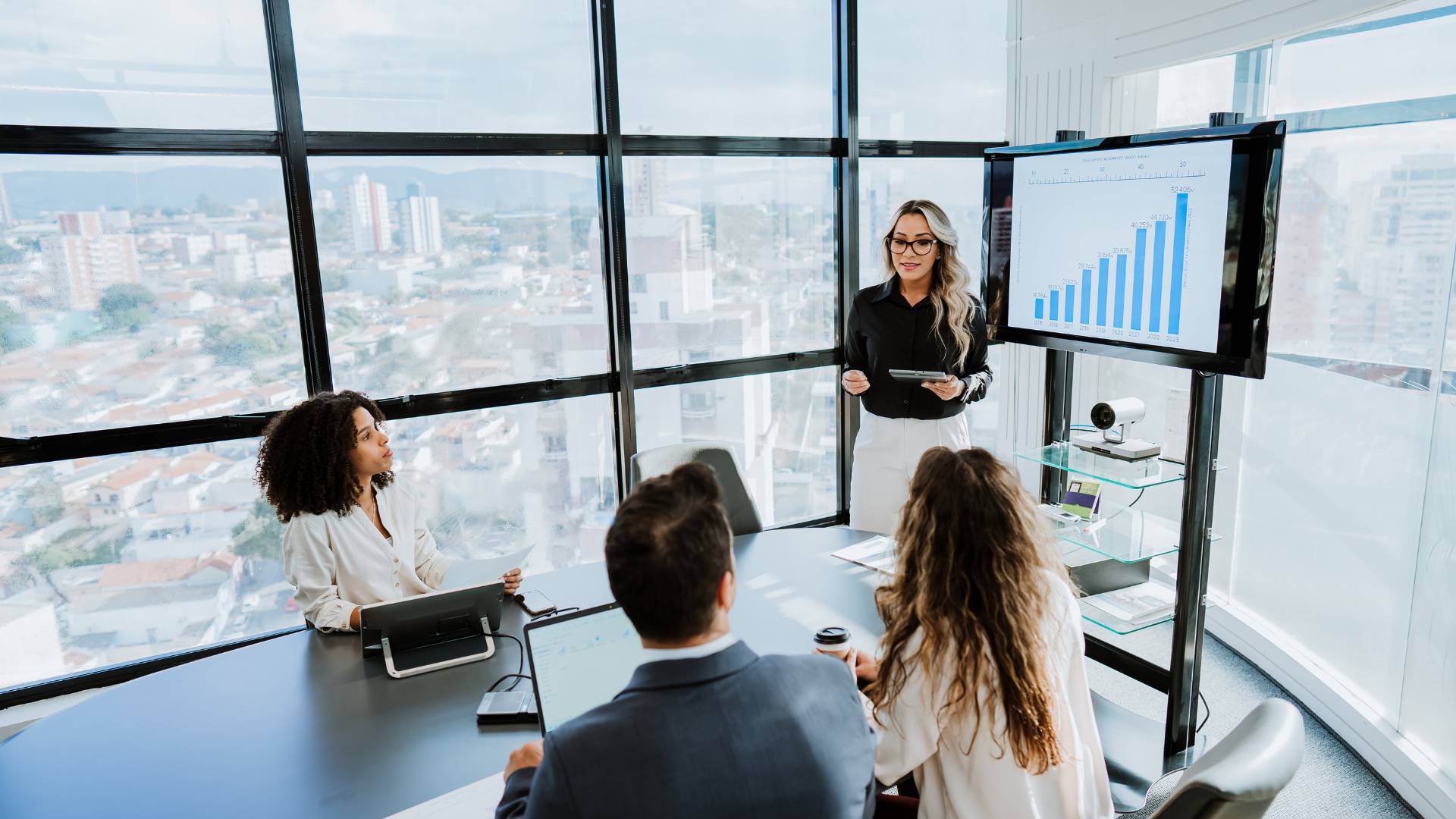 A team of four is having a meeting going over graphs that represent the health of the company. Three are seated and one woman is standing giving a presentation.