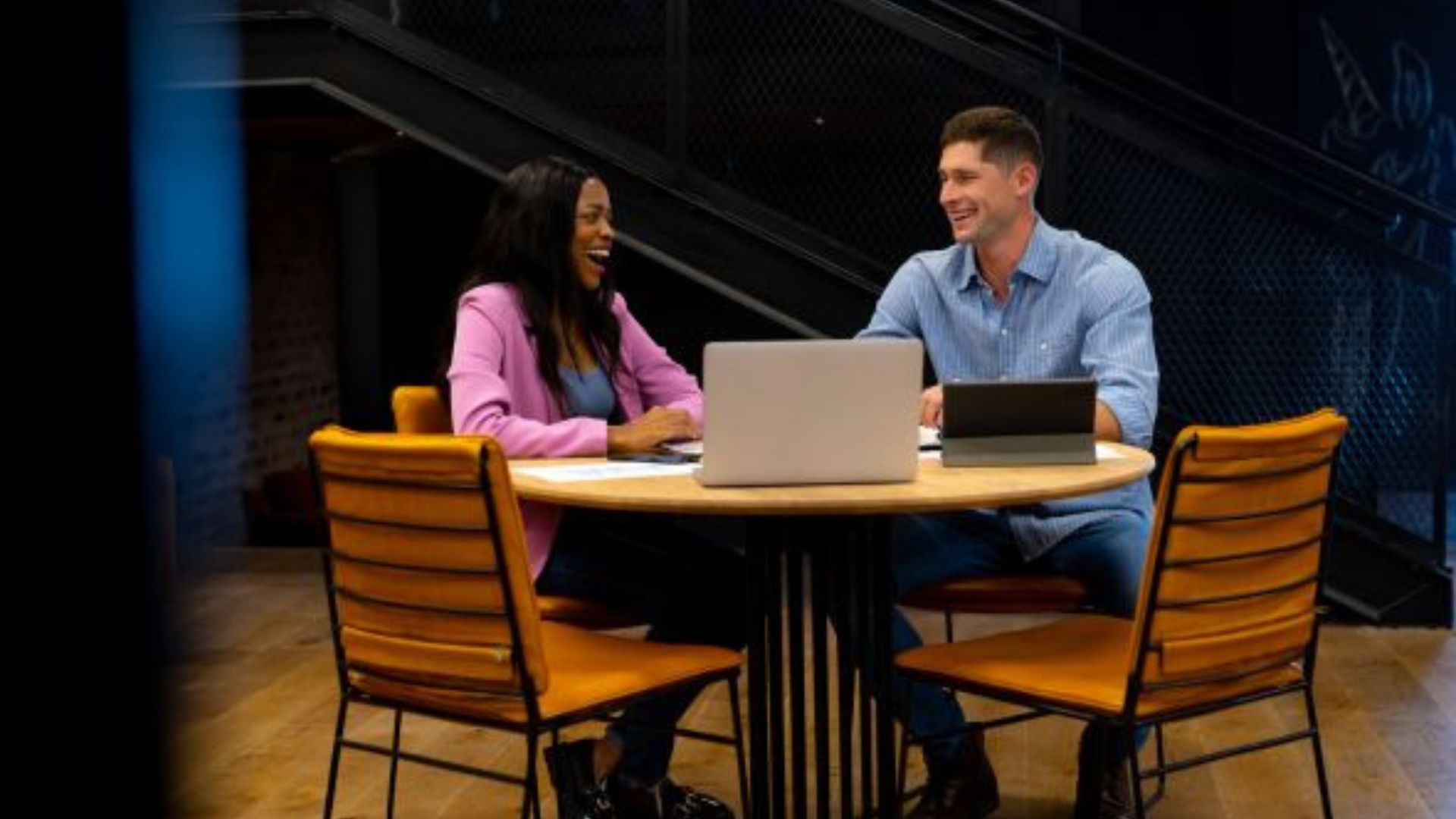 A man and woman sit at a round table during their quarterly one-on-one meeting. They’re smiling and engaging in conversation with a laptop open in front of each. They are in a casual office space with a stairway in the background, suggesting a relaxed but productive meeting environment.